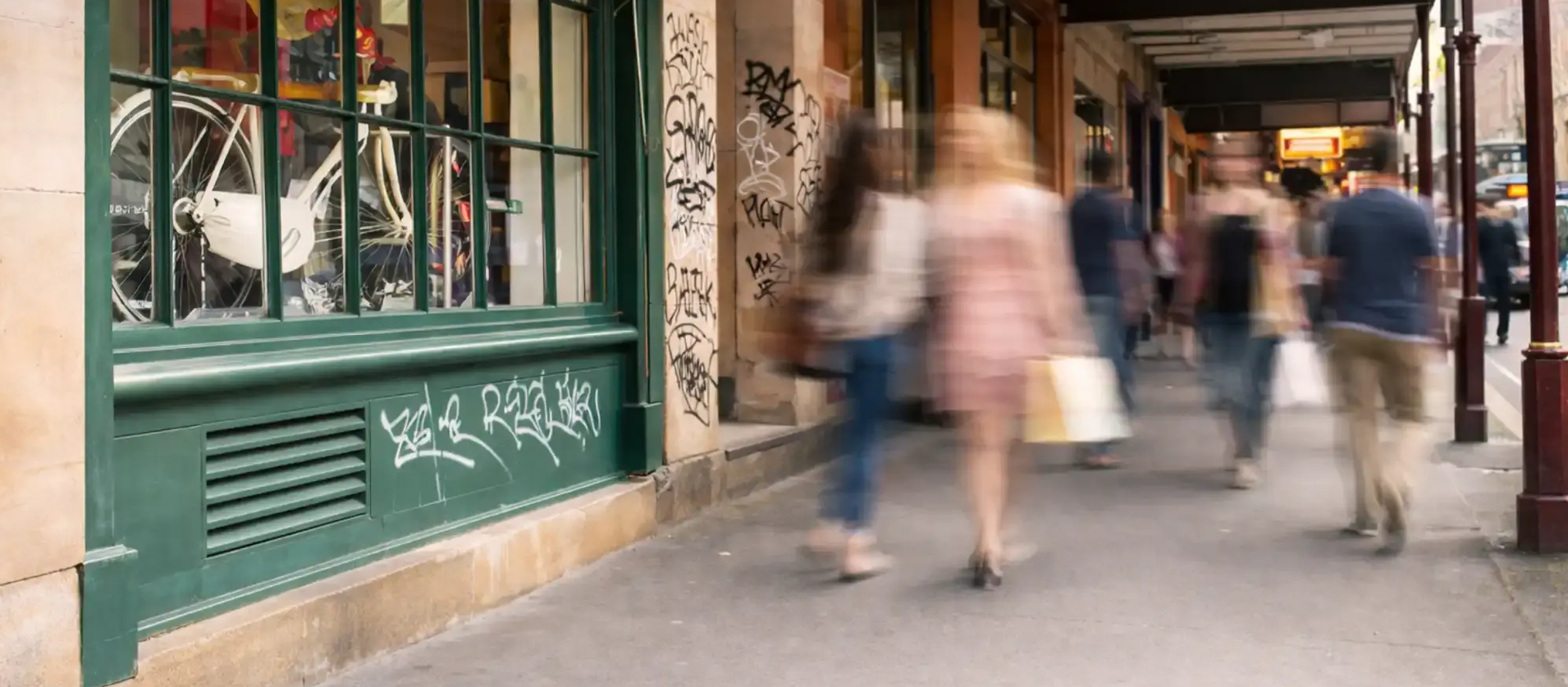 Busy commercial street with green-framed shopfront and sandstone columns marked with graffiti tagging, captured in long exposure so passing pedestrians appear as blurred, translucent figures.